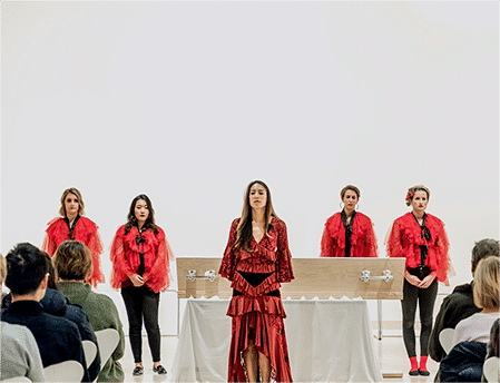 Five women in red clothes stand in front of a coffin on a white background with a crowd in front of them